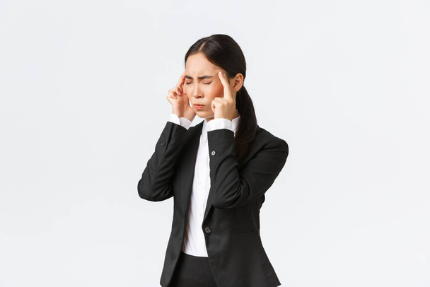 Profil einer asiatischen Geschäftsfrau im schwarzen Anzug mit Migräne, anrührenden Schläfen und Grimassen vor schmerzhaftem Gefühl im Kopf. Büroleiterin mit Kopfschmerzen, braucht Schmerzmittel, weißer Hintergrund - Foto, Bild
