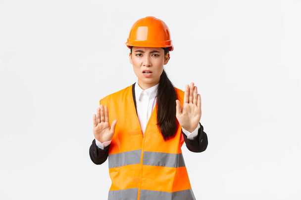Disappointed angry asian female engineer in safety helmet and reflective clothing saying stop, prohibit and disagree with construction manager, showing enough, no gesture, white background - Fotografie, Obrázek