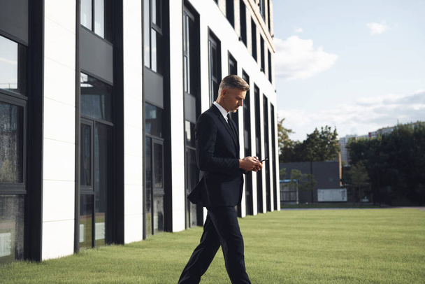 Confident mature businessman holding mobile phone while walking near office building - 写真・画像