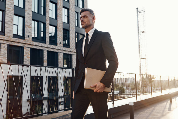 Sebevědomý dospělý muž v plném obleku s notebookem při chůzi před kancelářskou budovou - Fotografie, Obrázek