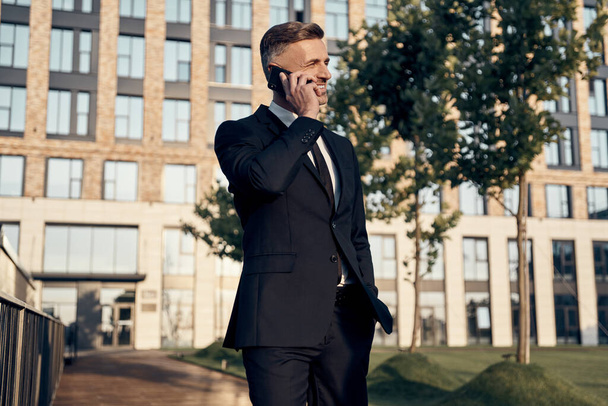 Впевнений зрілий бізнесмен розмовляє по мобільному телефону під час ходьби біля офісної будівлі на відкритому повітрі
 - Фото, зображення