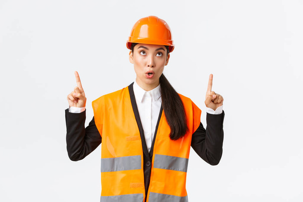 Building, construction and industrial concept. Surprised and curious asian female construction manager, engineer in safety helmet and reflective clothing, pointing fingers up at interesting banner - Photo, Image
