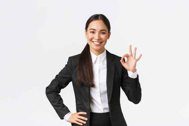 Gerente femenina profesional en traje negro buscando asertivo, animar todo bien, asegurar el trabajo hecho, mostrando gesto bien y sonriendo en aprobación, de pie satisfecho sobre fondo blanco
 - Foto, Imagen