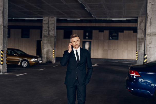 Confident mature businessman talking on mobile phone while walking by parking lot - Foto, Bild