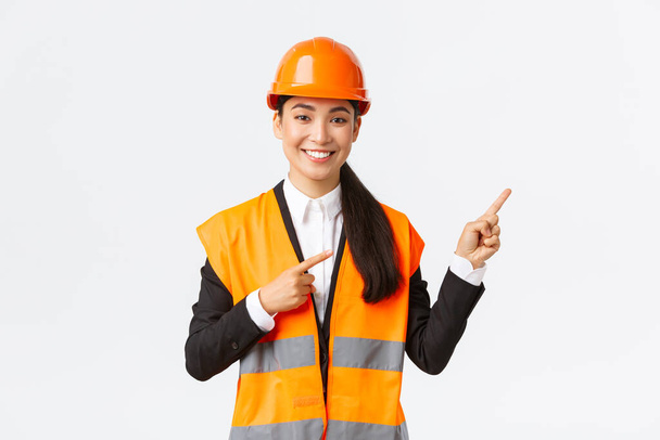 Épület, építőipar és ipari koncepció. Mosolygó női ázsiai mérnök biztonsági sisak és reflektív ruházat mutató tárgy, mutató ujjak jobb, eladó bemutatja birtok ügyfél - Fotó, kép