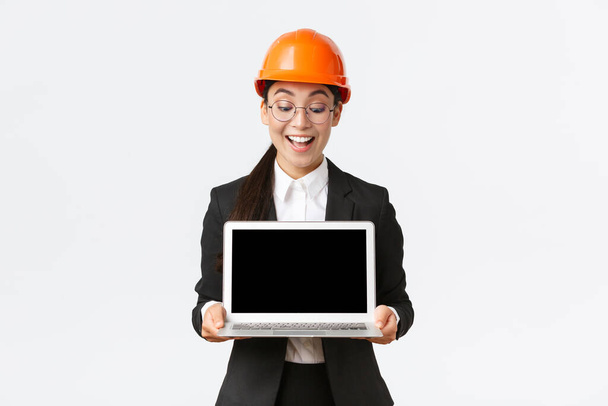 Impressed happy asian female chief engineer showing great results of enterprise to investors, standing in safety helmet and business suit, holding laptop and look pleased at computer screen - Foto, immagini