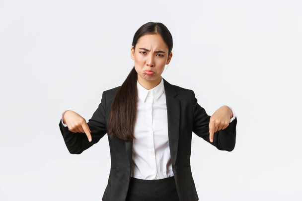 Disappointed sulking asian businesswoman complaining on unfair situation. Female entrepreneur in suit pointing fingers down and grimacing displeased, standing upset white background - Photo, Image