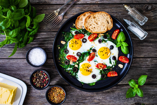 Sunny side up eggs with spinach, cherry tomatoes and black olives on wooden table  - 写真・画像