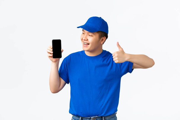 Teknoloji, online alışveriş ve nakliye konsepti. Mavi üniformalı, gülümseyen Asyalı teslimatçı başparmağını kaldırıyor ve cep telefonu ekranından memnun görünüyor. Tavsiye uygulaması, beyaz arka plan. - Fotoğraf, Görsel