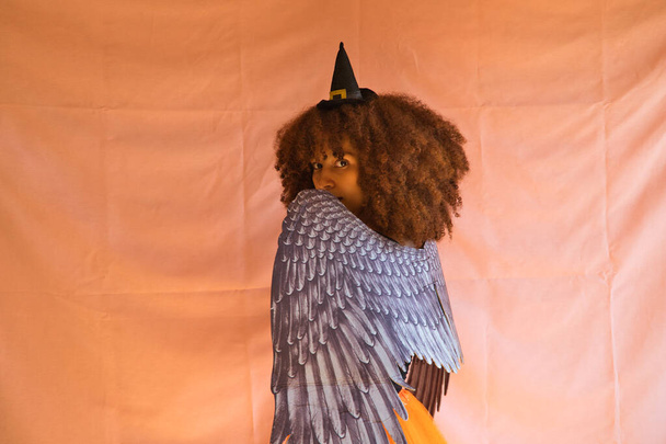 mladá Afroameričanka v kostýmu čarodějnice na Halloweenskou párty. Má na sobě čarodějnický klobouk, oranžovou sukni a křídla černého anděla. Žena se usmívá do kamery, zatímco dělá různé pózy. - Fotografie, Obrázek