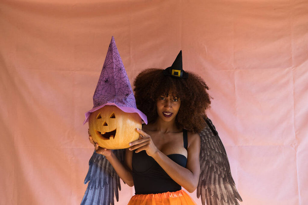jeune femme afro-américaine en costume de sorcière pour la fête d'Halloween. Elle porte un chapeau de sorcière, une jupe orange et des ailes d'ange noir. La femme sourit à la caméra tout en jouant avec la citrouille. - Photo, image