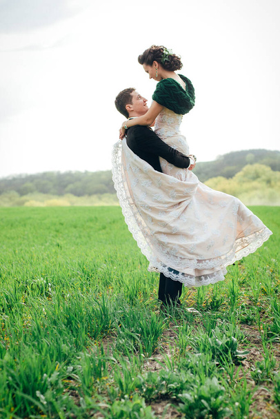 el novio en un traje marrón y la novia en un vestido de color marfil en un campo verde retrocediendo en la distancia contra el cielo - Foto, Imagen