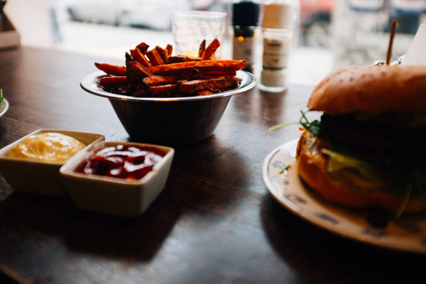 A high-angle shot of a wooden table with ketchup and cheddar sauces, burger and french fries - Photo, Image