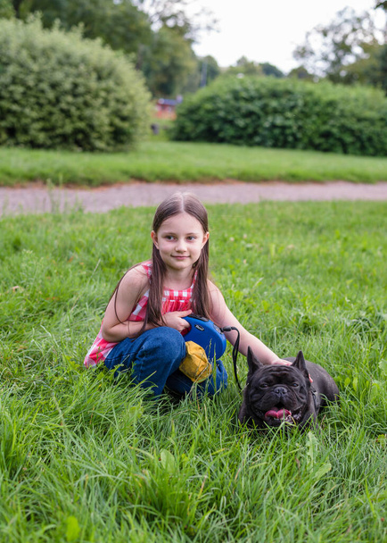 Una bambina di 6 anni cammina con un cane nel parco. Il Bulldog francese non vuole camminare, riposa e mente. La ragazza tira il cane al guinzaglio. - Foto, immagini