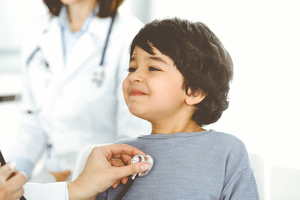 Doctor-woman examining a child patient by stethoscope. Cute arab boy at physician appointment. Medicine concept - 写真・画像