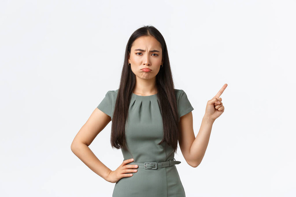 Majitelé malých podniků, podnikatelky. Skeptická a zklamaná asijská žena v šatech stěžující si, ukazující prstem v pravém horním rohu na šklebu, trucující, bílé pozadí - Fotografie, Obrázek