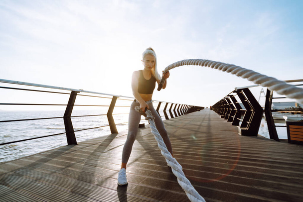 Young woman wearing sports clothes is doing exercises at the beach pier in the morning. Battle ropes. Kettlebell. Sport, Active life. - Foto, Bild