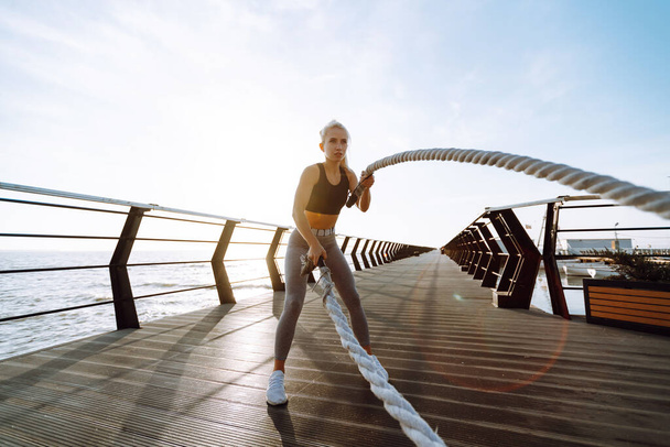 Young woman wearing sports clothes is doing exercises at the beach pier in the morning. Battle ropes. Kettlebell. Sport, Active life. - Foto, Imagem