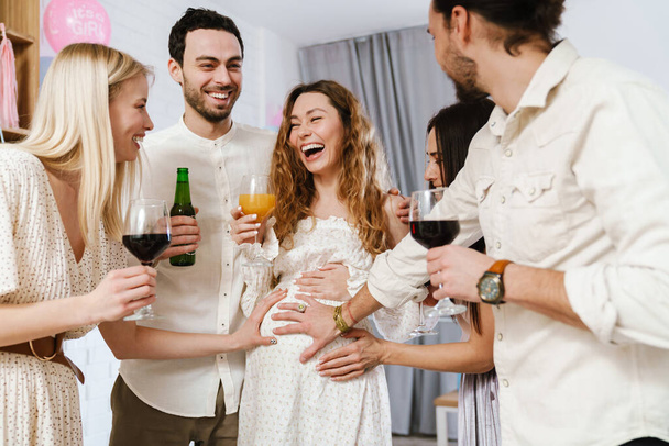 Young happy pregnant woman making fun with her friends during gender reveal party indoors - Фото, изображение