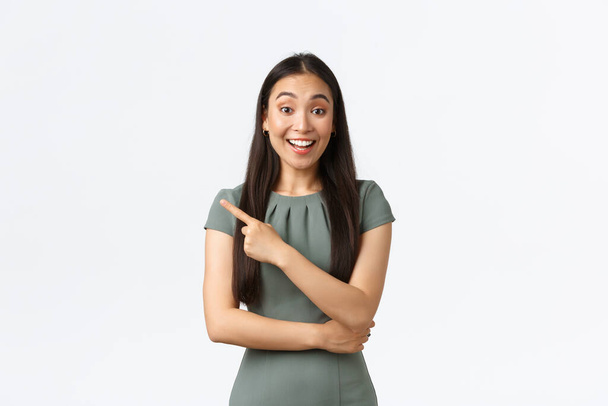 Proprietários de pequenas empresas, conceito de mulheres empresárias. excitado feliz asiático fêmea no vestido sorrindo e apontando dedo esquerdo no anúncio, olhando espantado, branco fundo
 - Foto, Imagem