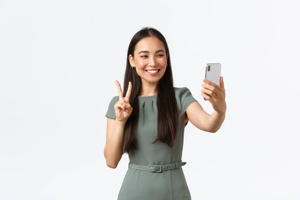 Kleinunternehmer, Unternehmerinnen. Wunderschöne weibliche asiatische Bloggerin macht Selfie auf dem Smartphone, posiert und schaut in die Handykamera mit Friedenszeichen - Foto, Bild