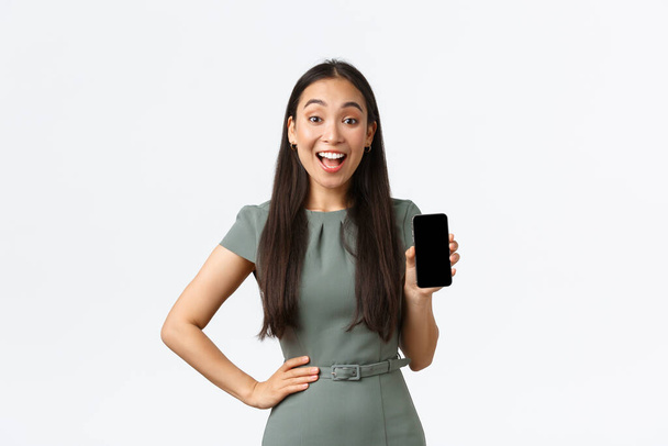 Kleinunternehmer, Unternehmerinnen. Aufgeregt asiatische Geschäftsfrau macht Ankündigung, zeigt Smartphone-Bildschirm Anwendung mit erstaunten Lächeln, weißer Hintergrund - Foto, Bild