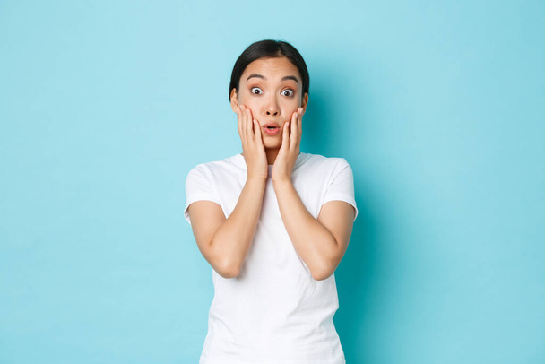 Surprised and amazed asian female student react to amazing news, looking impressed, gasping and saying wow impressed, standing light-blue background astounded - Photo, Image