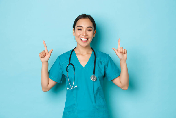 Covid-19, social distancing and coronavirus pandemic concept. Smiling attractive asian female nurse, doctor or intern in scrubs, pointing fingers up, telling big news, light blue background - Photo, Image