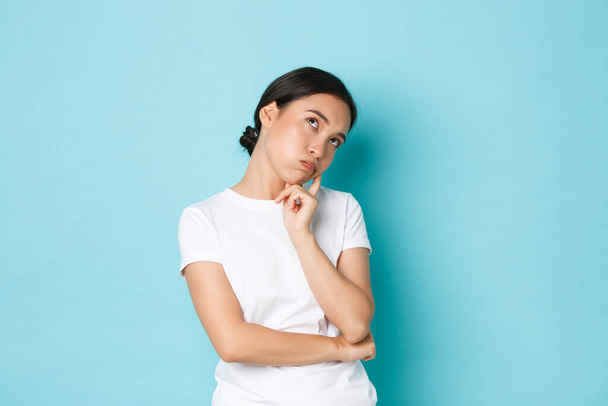Bored or annoyed cute asian girl in white t-shirt roll eyes, lean on palm and looking indifferent, being not in mood, feeling boredom and apathy, standing blue background not concerned - Foto, afbeelding