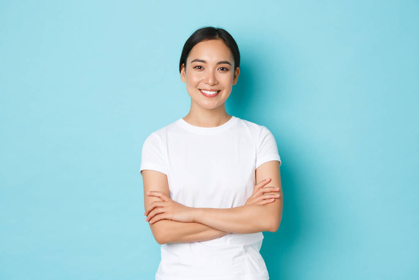 Porträt eines selbstbewussten asiatischen Mädchens, das zufrieden lächelt, Brust mit verschränkten Armen, selbstbewusste Pose, Studentin, die optimistisch und entschlossen wirkt, blauer Hintergrund, lässige Kleidung, Lifestylekonzept - Foto, Bild