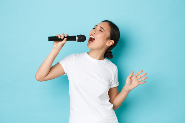Styl życia, ludzie i pojęcie czasu wolnego. Namiętna i beztroska ładna azjatycka dziewczyna śpiewająca piosenkę w mikrofonie, zginająca się podczas występu, jak karaoke, stojące jasnoniebieskie tło wykonujące - Zdjęcie, obraz