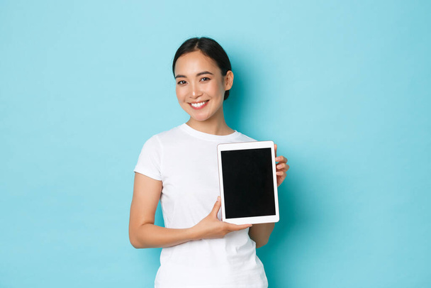 Portrait de sourire attrayant asiatique fille démontrer le projet ou la publicité sur l'écran de la tablette numérique, introduire un nouveau site d'achat, debout optimiste sur fond bleu clair - Photo, image