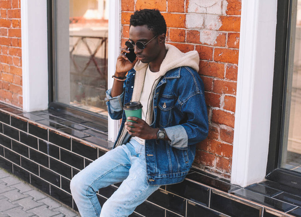Portrait of young african man talking on smartphone while sitting with cup of coffee on a city street - Fotografie, Obrázek