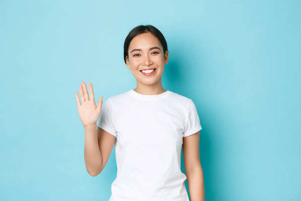 Portrait of friendly cheerful asian girl in white t-shirt saying hello, greeting people, smiling and waving hand in hi gesture, standing blue background upbeat, welcome someone - Photo, Image