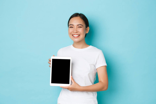 Portrait de fière et heureuse fille asiatique souriante, souriant joyeusement, regardant heureux tout en montrant l'écran de la tablette numérique, démontrer son projet de travail ou de publicité, fond bleu clair - Photo, image