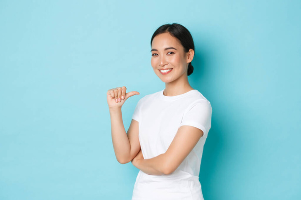 Уверенная улыбающаяся азиатка в белой футболке, указывающая на себя с гордостью, напористостью, хвастовством, продвижением собственных способностей, профессионализмом, поиском работы, стоящим на синем фоне - Фото, изображение