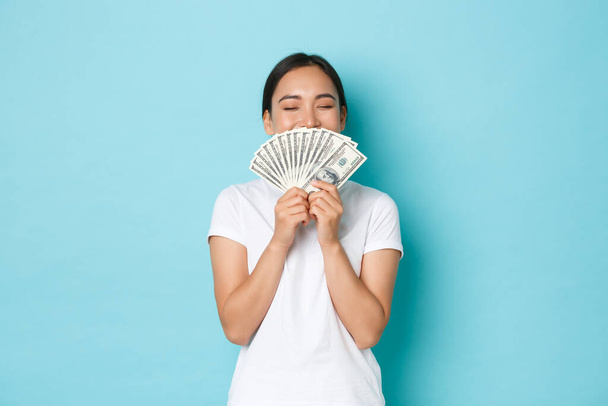 Shopping, money and finance concept. Young successful and lucky cheerful asian girl smelling dollars and smiling satisfied, enjoying being rich, holding cash over light-blue background - Fotó, kép