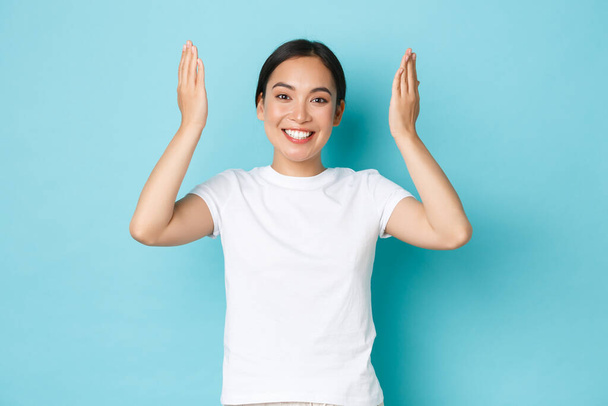 Cheerful happy asian girl open eyes and smiling cheerful and surprised, raising hands up and looking upbeat over blue background, celebrating birthday, being amazed with gift - Photo, Image