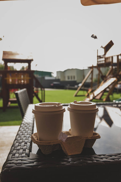 Zwei weiße Pappbecher auf dem Tisch vor dem Hintergrund eines verschwommenen Spielplatzes. ein Imbiss an der frischen Luft. Selektiver Fokus - Foto, Bild