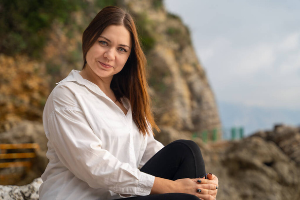 Happy young brunette woman in white shirt walks around resort town and laughs, posing against mountains. The calm before the storm. Idyllic walk alone - Zdjęcie, obraz