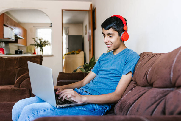 Hispanic teenage boy with headphones listening to music while using laptop at home in Latin America - Fotó, kép