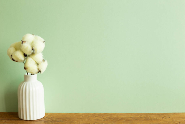 Белая ваза хлопкового растения на деревянном столе. зеленый фон стены. Интерьер дома - Фото, изображение