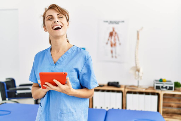 Joven fisioterapeuta caucásica chica sonriendo feliz usando touchpad en la clínica. - Foto, imagen