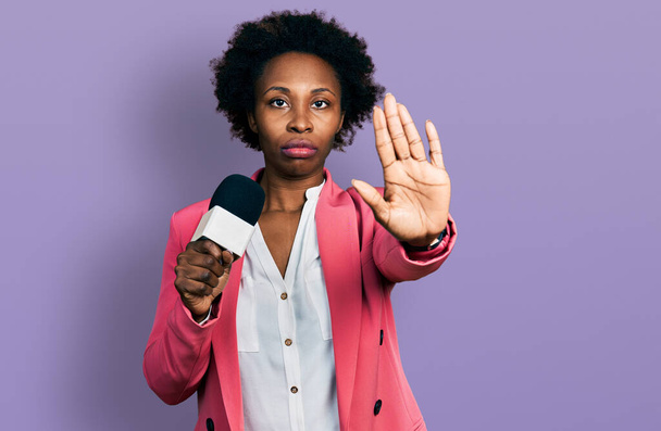 Αφρο-αμερικανική γυναίκα με αφρο μαλλιά κρατώντας το μικρόφωνο δημοσιογράφος με ανοιχτό χέρι κάνει στοπ υπογράψει με σοβαρή και σίγουρη έκφραση, χειρονομία άμυνας  - Φωτογραφία, εικόνα