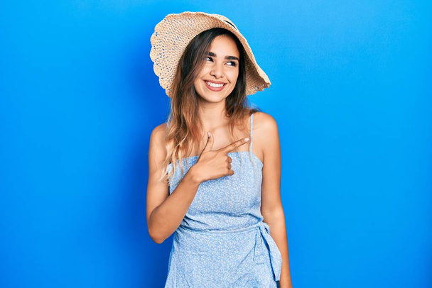 Young hispanic girl wearing summer hat cheerful with a smile of face pointing with hand and finger up to the side with happy and natural expression on face  - Foto, afbeelding