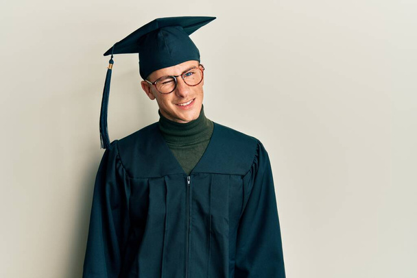 Young caucasian man wearing graduation cap and ceremony robe winking looking at the camera with sexy expression, cheerful and happy face.  - Foto, imagen
