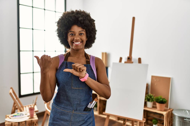 Joven mujer afroamericana con cabello afro en el estudio de arte apuntando hacia atrás con la mano y los pulgares hacia arriba, sonriendo confiada  - Foto, imagen