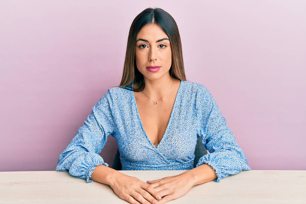 Jeune femme hispanique portant des vêtements décontractés assis sur la table avec une expression sérieuse sur le visage. simple et naturel regardant la caméra.  - Photo, image