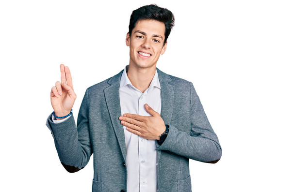 Jovem hispânico vestindo roupas de negócios sorrindo jurando com a mão no peito e os dedos para cima, fazendo uma promessa de lealdade juramento  - Foto, Imagem
