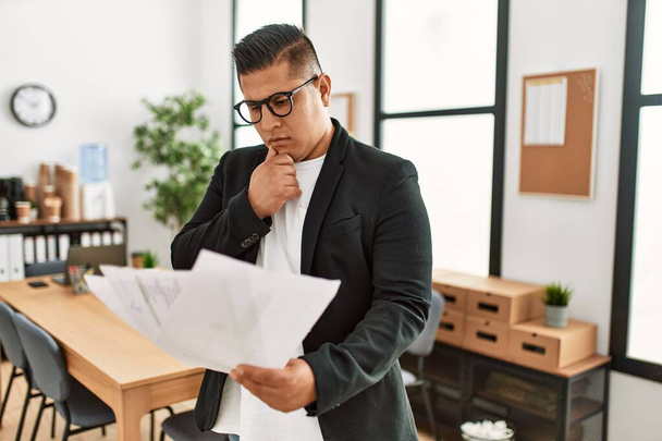 Молодой латинский бизнесмен с серьезным выражением лица читает бумажки стоя в офисе. - Фото, изображение